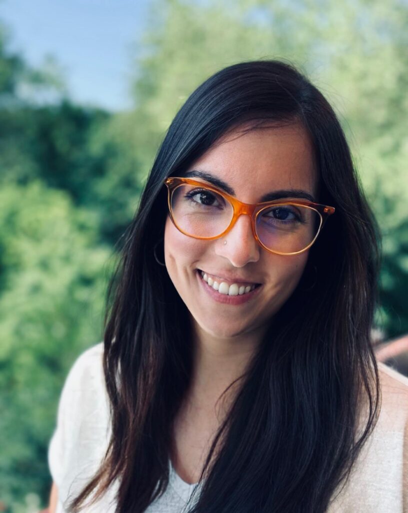 Eine lächelnde Frau mit Brille vor einem grünen Hintergrund mit Bäumen in Großaufnahme